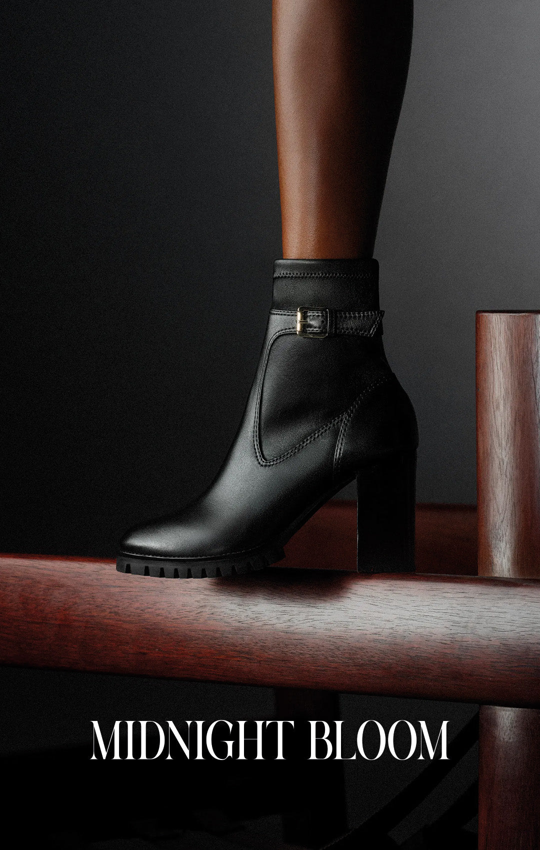  Black leather ankle boot with a high block heel rests on a wooden rail against a dark background; “MIDNIGHT BLOOM” text below, in a moody fashion ad. Shop Now.