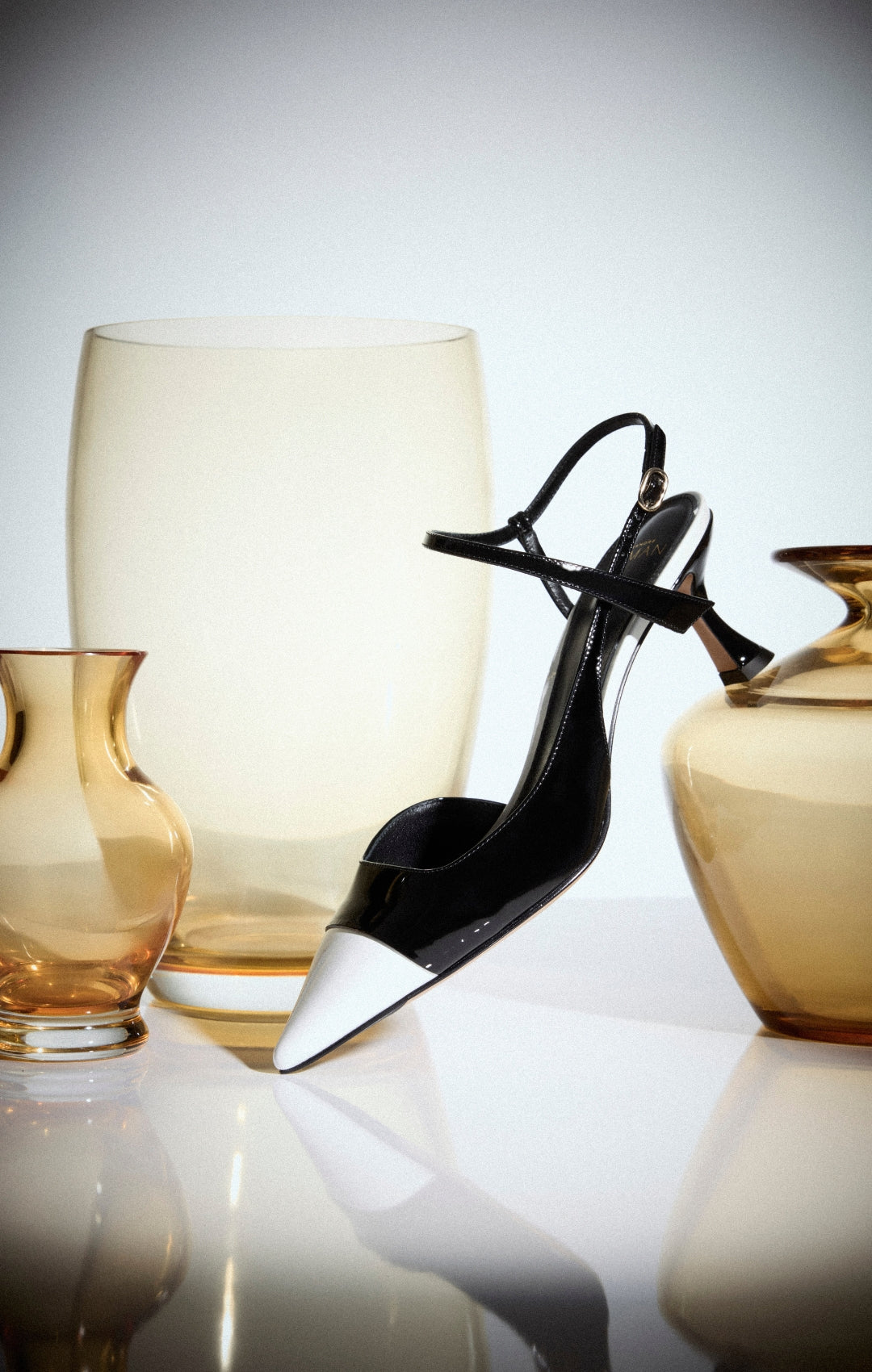  Black-and-white scissors stand open on a glossy surface, framed by amber glass vessels against a pale, minimalist background.Shop Now.