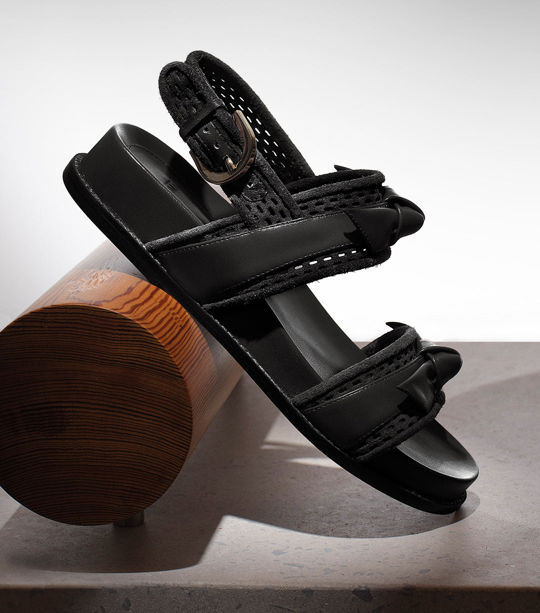 Black sport sandal with laser-cut detailing is displayed resting on a wooden cylinder against a neutral background. White text below reads “NEW FLATS.”