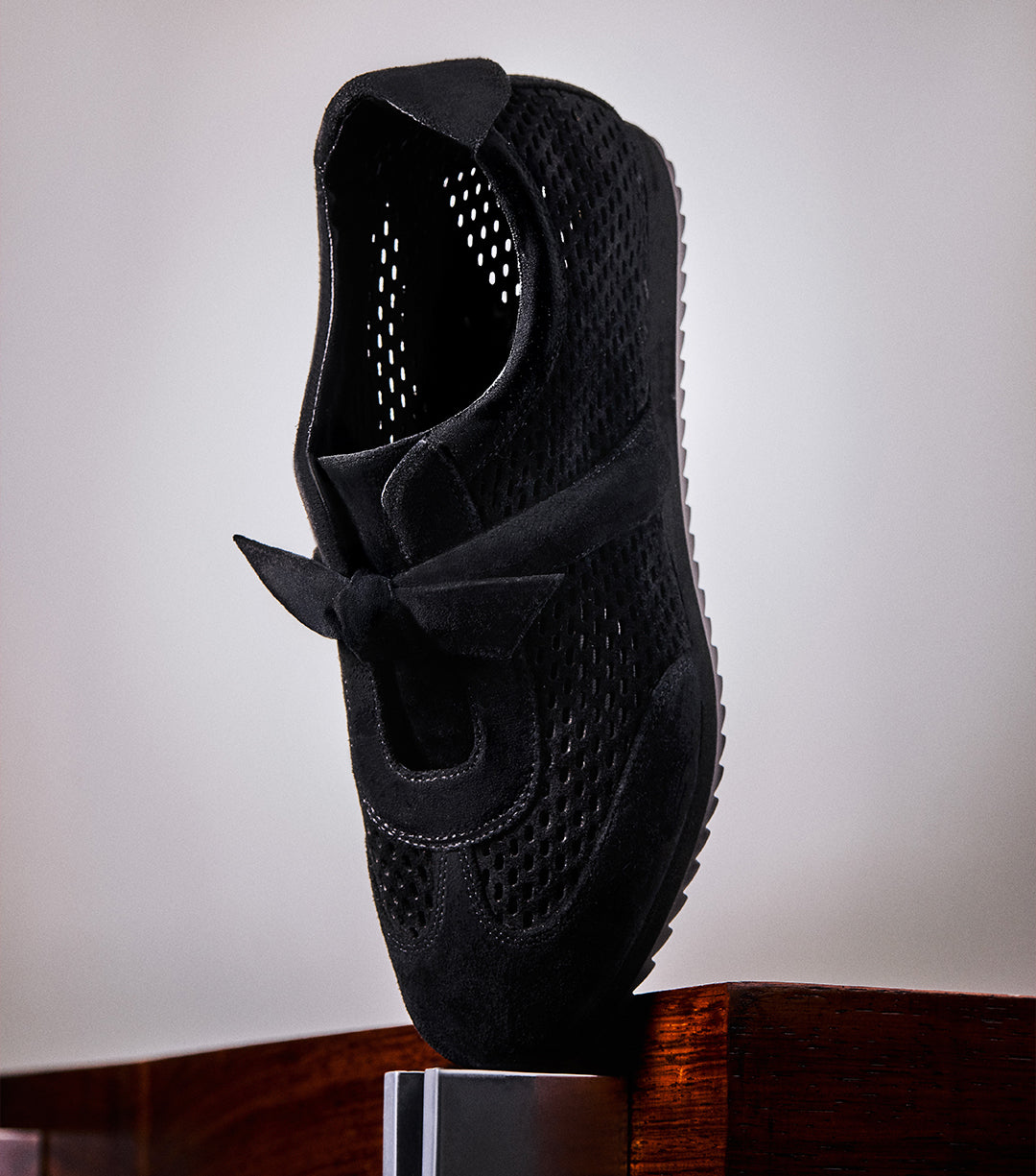 A black sneaker with laser-cut detailing and a bow accent is displayed balanced on a wooden surface against a neutral background. White text below reads “NEW SNEAKERS.”