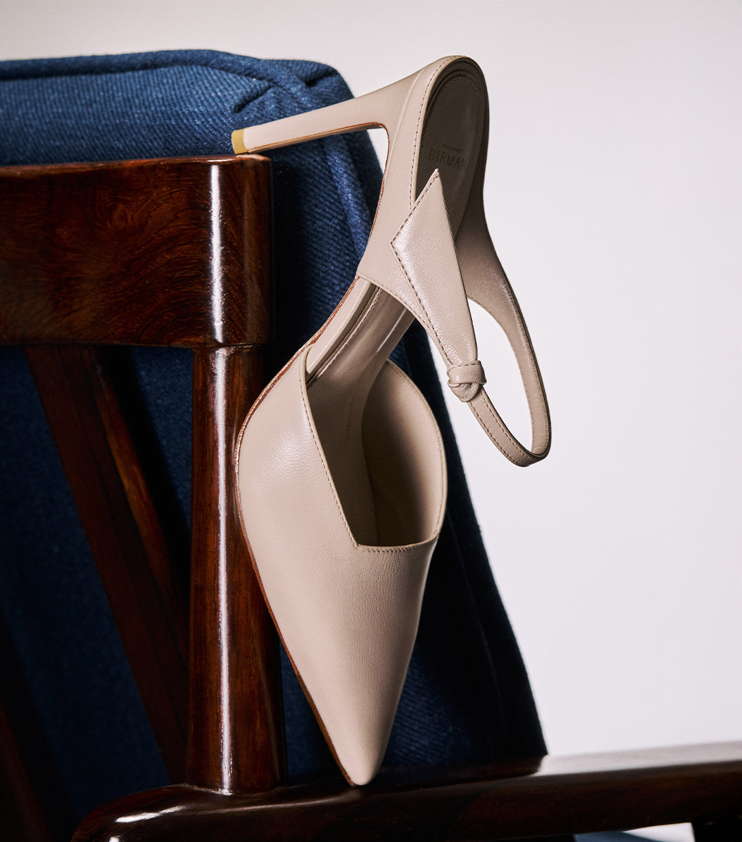 Beige pump displayed elegantly on the edge of a dark wooden chair, with a blue fabric on the background. Text on image reads "New Pumps".