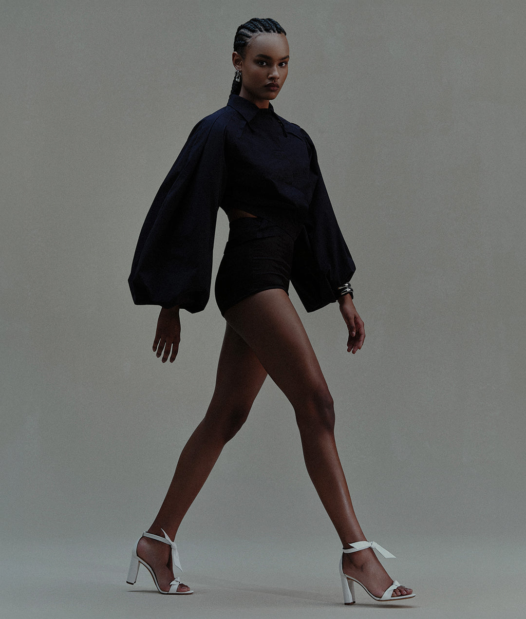 A woman with braided hair walking, wearing a dark voluminous top, black shorts, and white block-heel sandals with white ribbon ankle ties.
