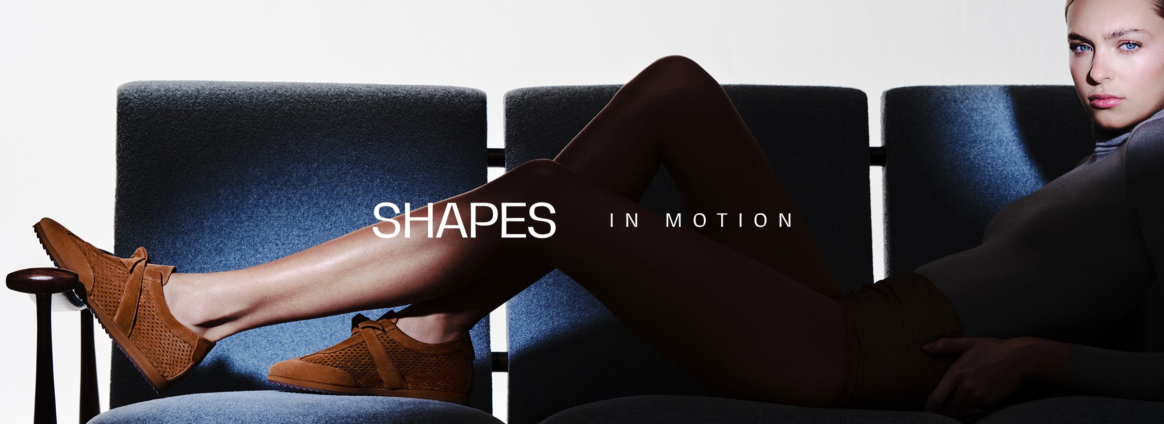  A woman sitting on a dark chair wearing brown suede perforated sneakers. Text overlay: SHAPES IN MOTION.