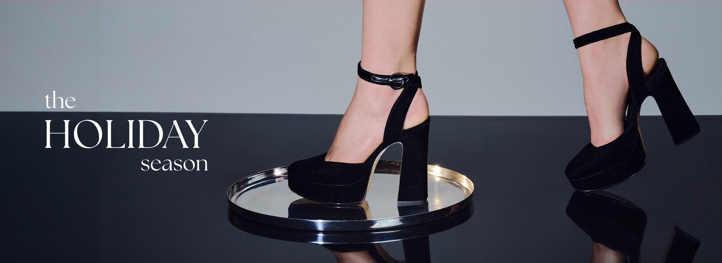  close-up of a person's legs wearing black suede platform high heels with thick block heels and ankle straps. One heel is resting on a mirrored silver tray. Text overlay: the HOLIDAY season.