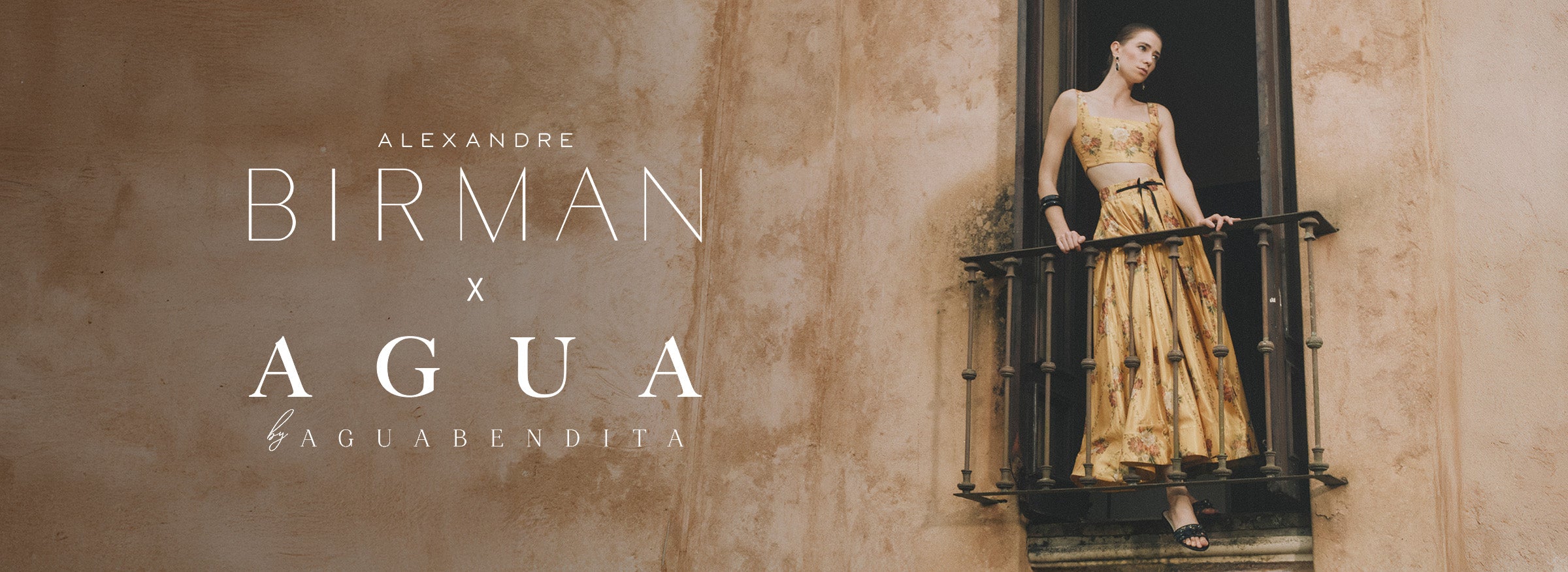  A campaign image featuring a model standing on a balcony with an iron railing, wearing a yellow two-piece set with a floral print. White text overlay in the center reads: "ALEXANDRE BIRMAN x AGUA by AGUA BENDITA”.