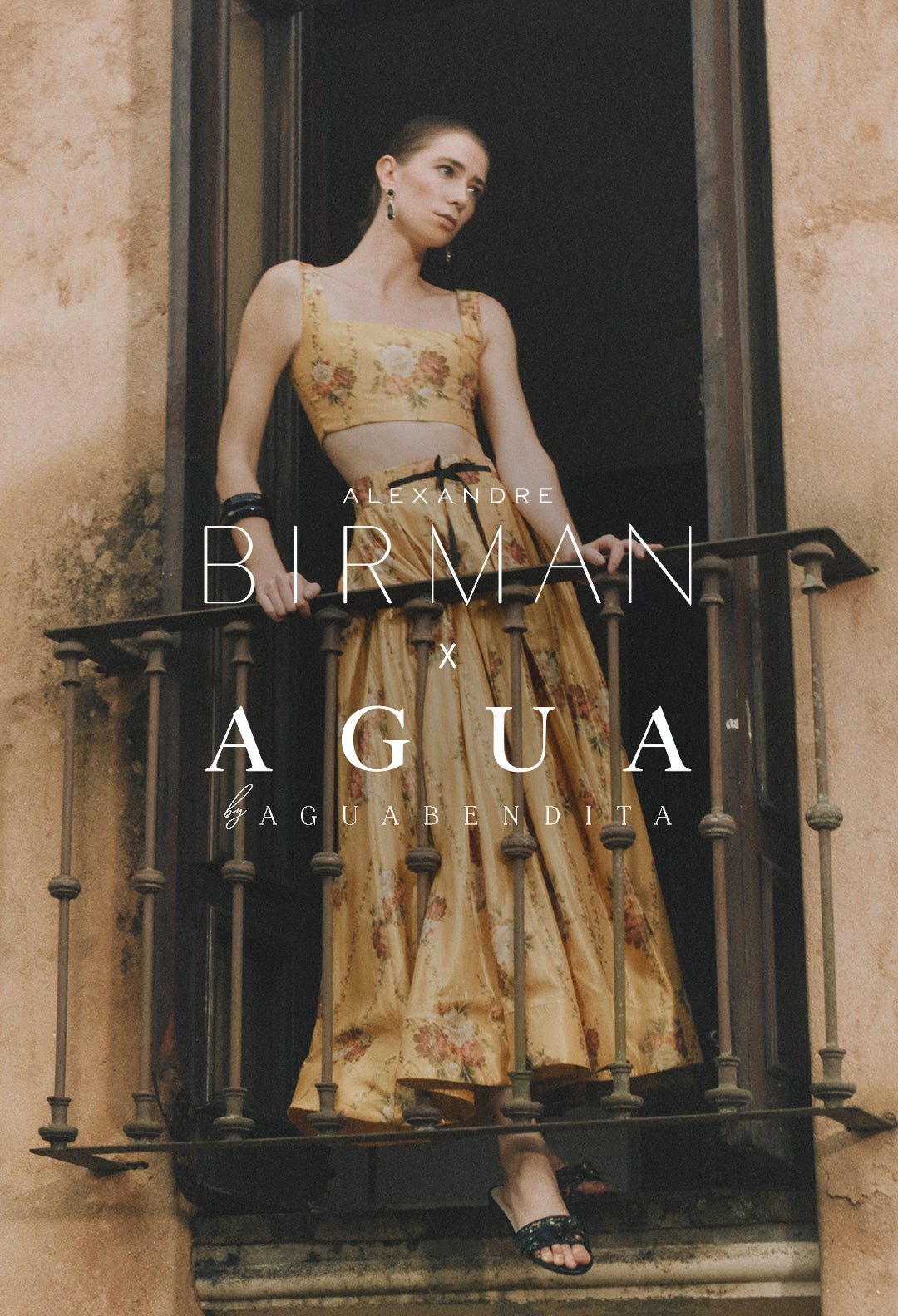  A campaign image featuring a model standing on a balcony with an iron railing, wearing a yellow two-piece set with a floral print. White text overlay in the center reads: "ALEXANDRE BIRMAN x AGUA by AGUA BENDITA”.