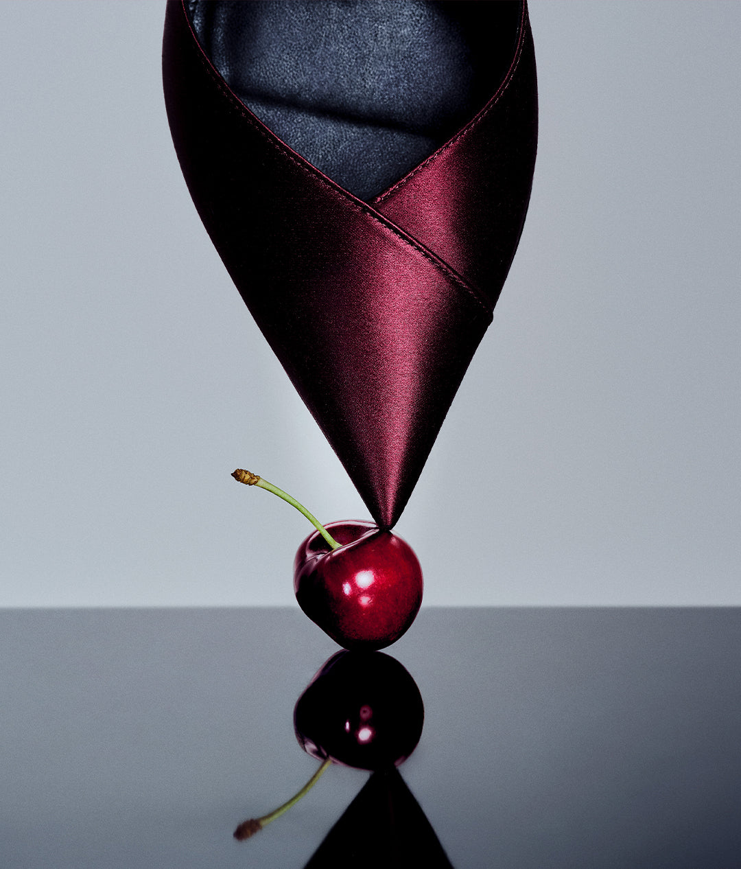  An artistic close-up showing the pointed toe of a burgundy satin shoe balancing vertically on top of a single red cherry. The composition is mirrored on a sleek, reflective black surface.