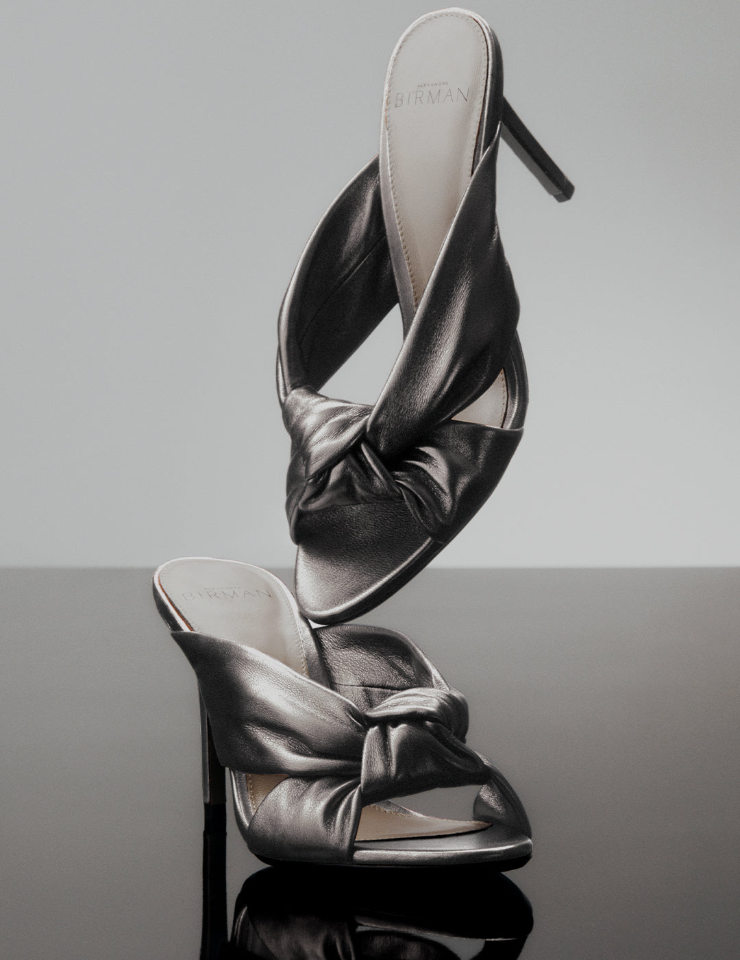 A shiny metallic pink high-heel slide sandal with two thin straps, set against a bright orange circular background element.