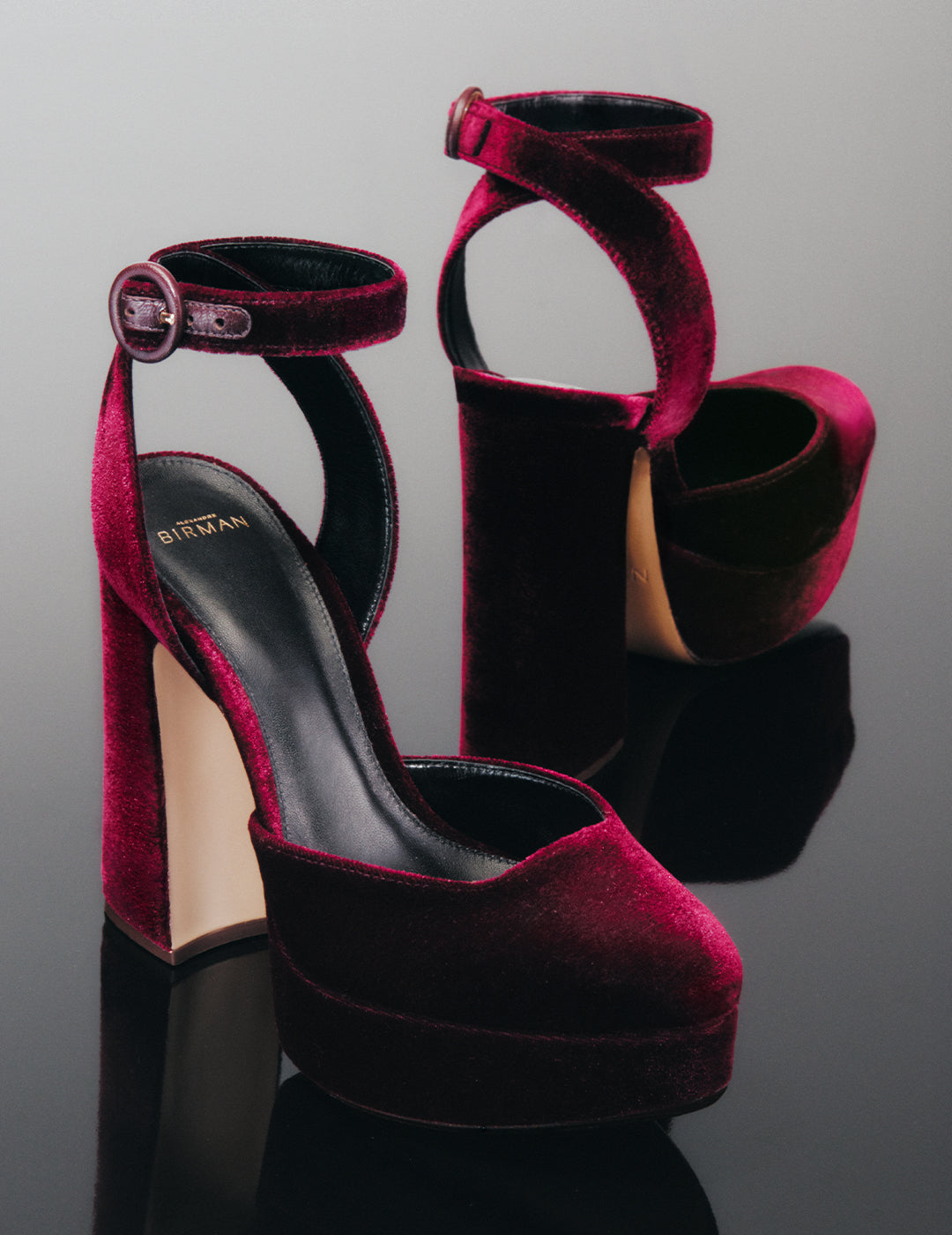 Deep burgundy velvet platform pumps displayed on a reflective surface. The shoes feature a chunky high heel, a thick platform sole, and a secure ankle strap with a buckle.