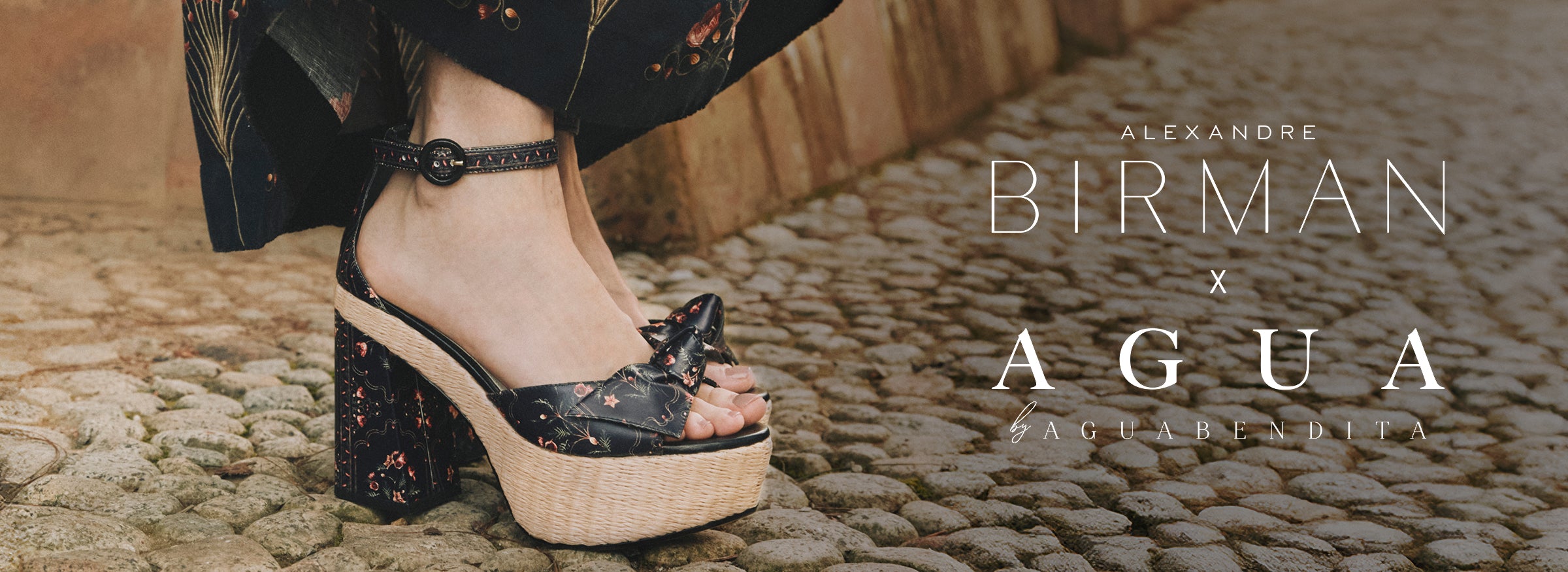  A model wearing a strapless dark floral dress standing outdoors next to a stone fountain with lily pads, featuring the text overlay "Alexandre Birman x Agua by Agua Bendita - Shop Now."