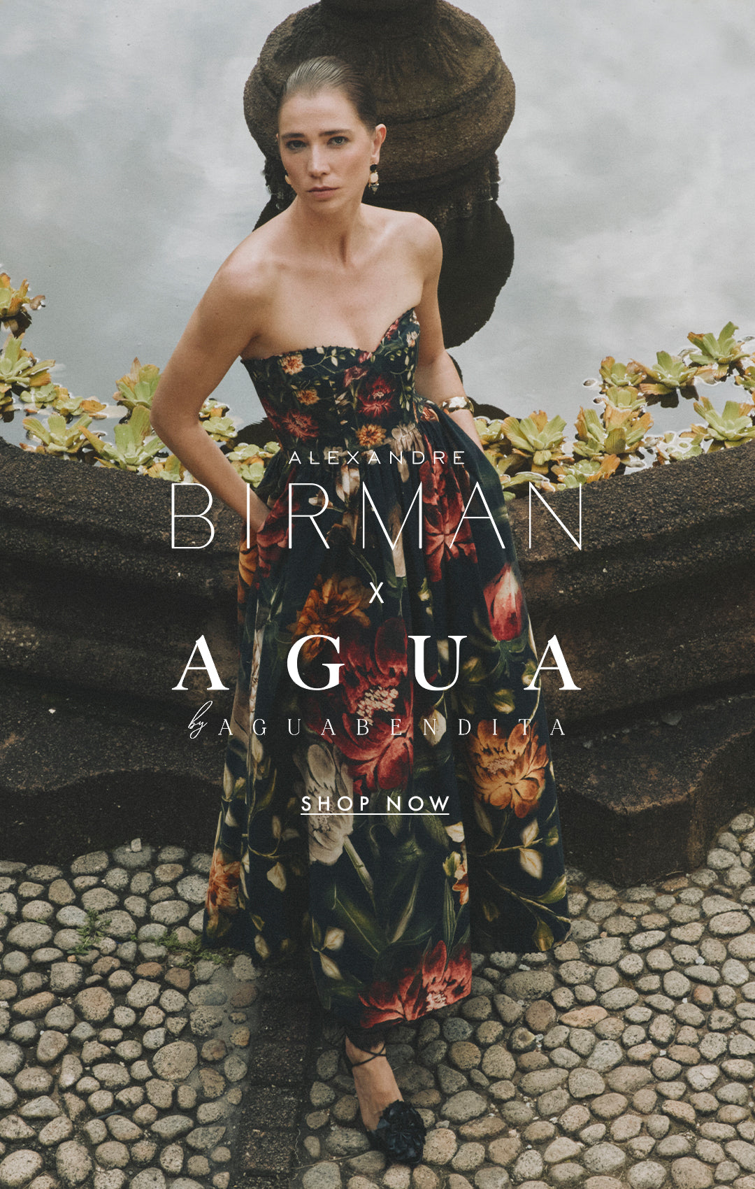  A model wearing a strapless dark floral dress standing outdoors next to a stone fountain with lily pads, featuring the text overlay "Alexandre Birman x Agua by Agua Bendita - Shop Now."
