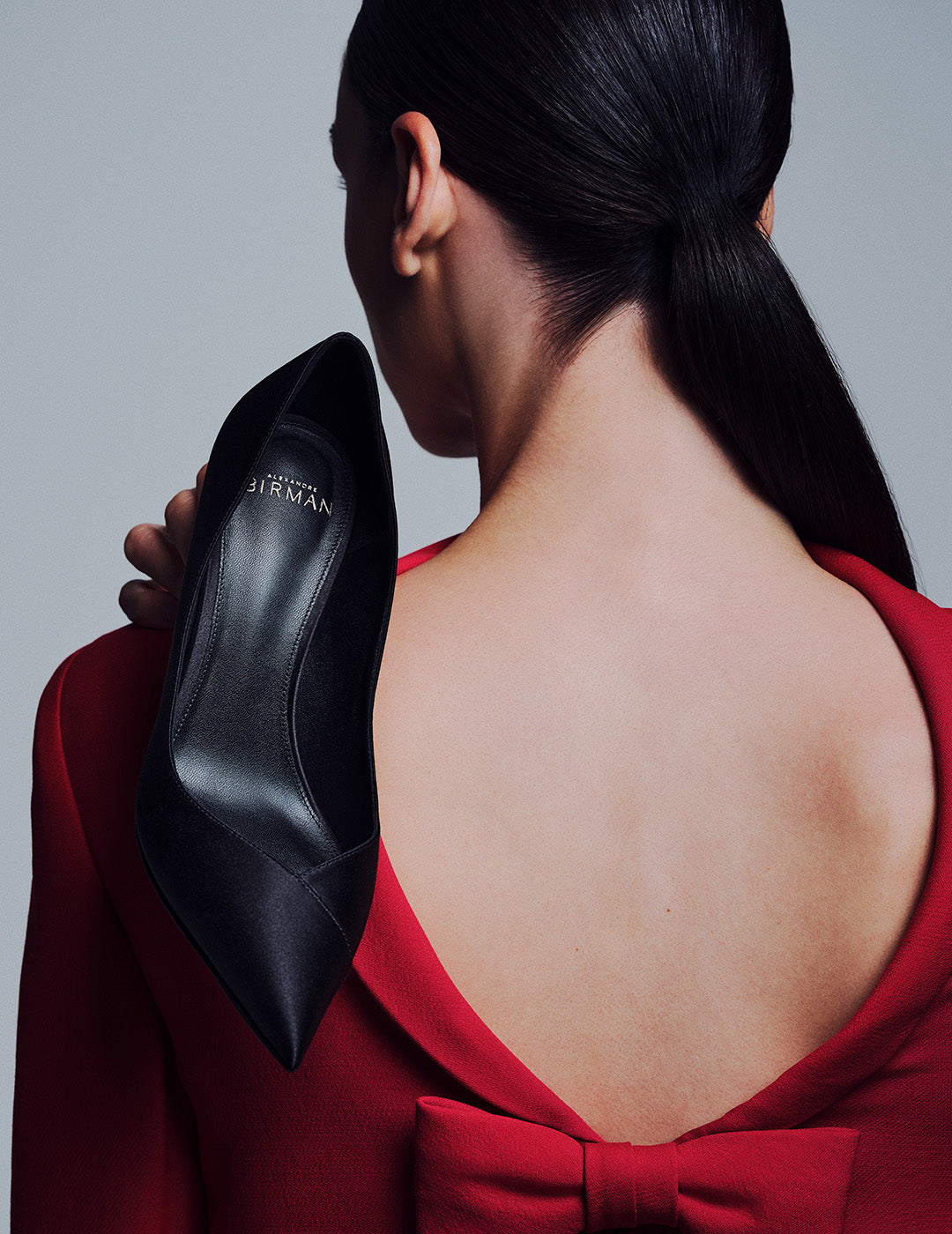 The rear view of a woman wearing a red dress with a bow detail, holding a black pointed-toe pump over her shoulder, revealing the Alexandre Birman logo on the insole.