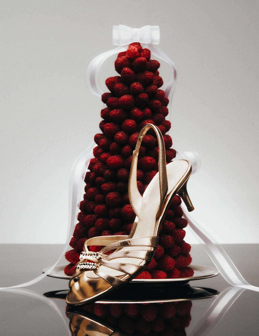 A metallic gold heeled sandal with crystal embellishments displayed in front of a festive, cone-shaped tower made of red raspberries topped with a white bow and ribbon.