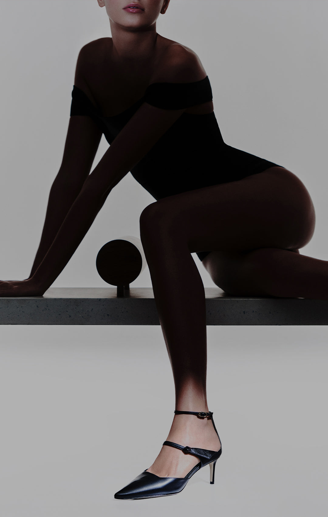  A moody, silhouetted shot of a model sitting on a minimalist bench, wearing a black bodysuit. The focus is on her legs and footwear: a pair of sleek, black pointed-toe pumps with delicate ankle straps and a kitten heel.