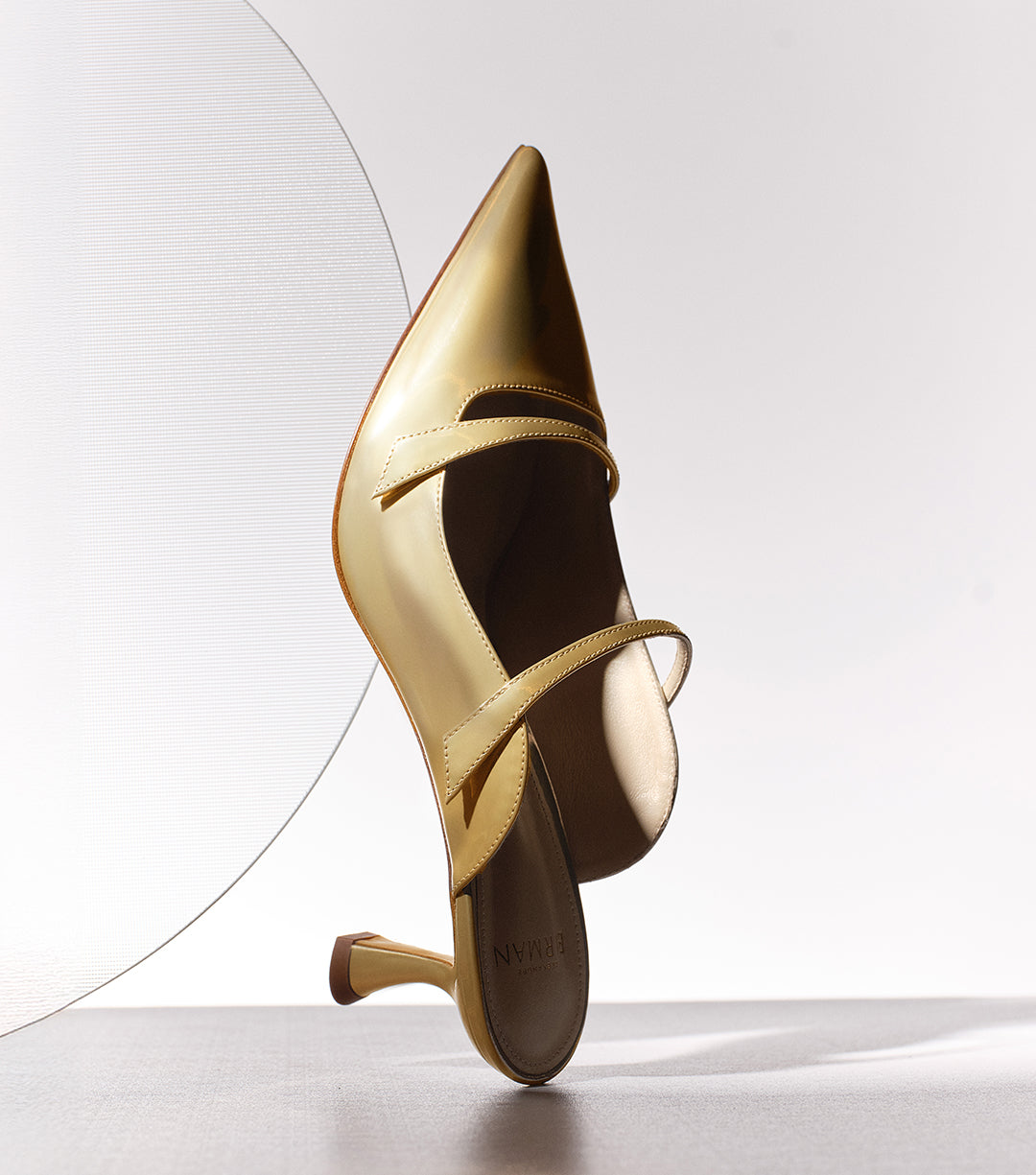 A yellow patent leather pointed-toe mule with a sculptural kitten heel and instep straps, balanced upright against a white background with a sheer textured circular prop.