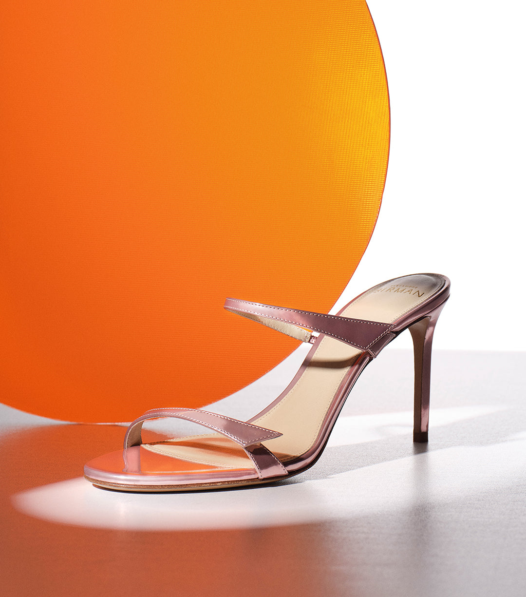 A metallic pink high-heeled mule sandal with thin straps, displayed in front of a bright orange translucent circular prop against a white background.