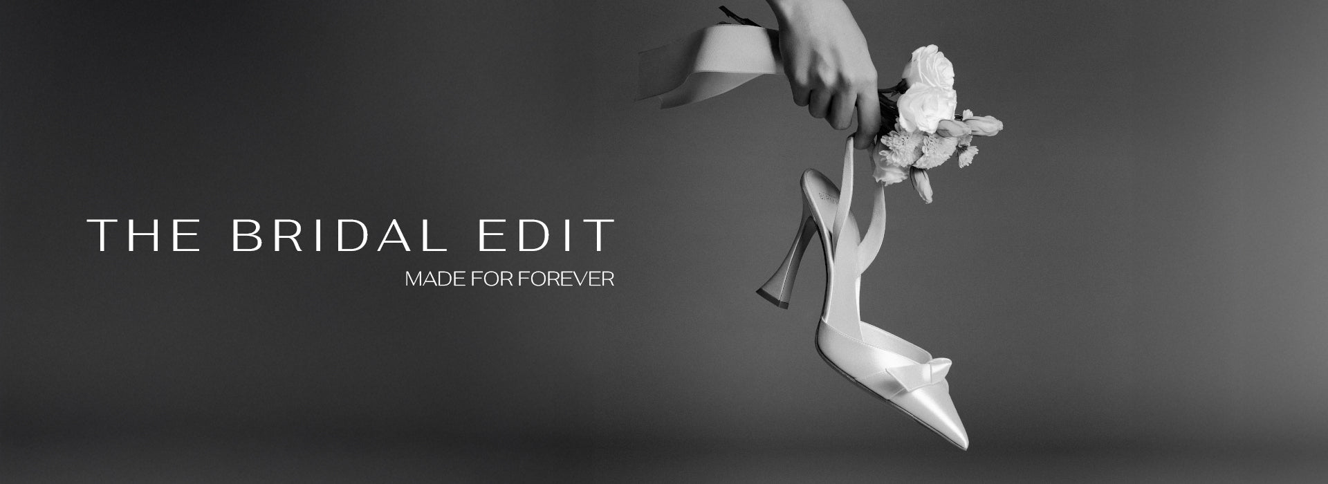  Grayscale image of a bridal shoe with ribbon and floral bouquet held against a blurred background. Text reads: "The Bridal Edit, Made for Forever," conveying elegance. Tap to shop