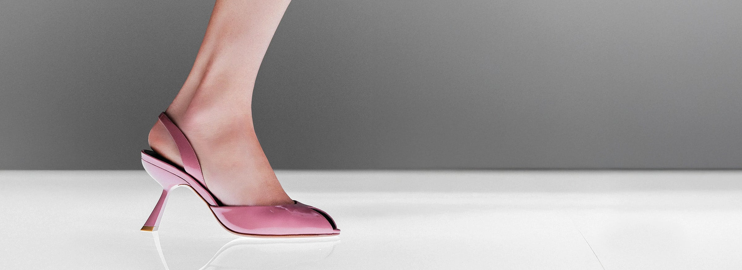  Model wearing a pink patent leather peep-toe slingback pump with a sculptural heel against a gray background.