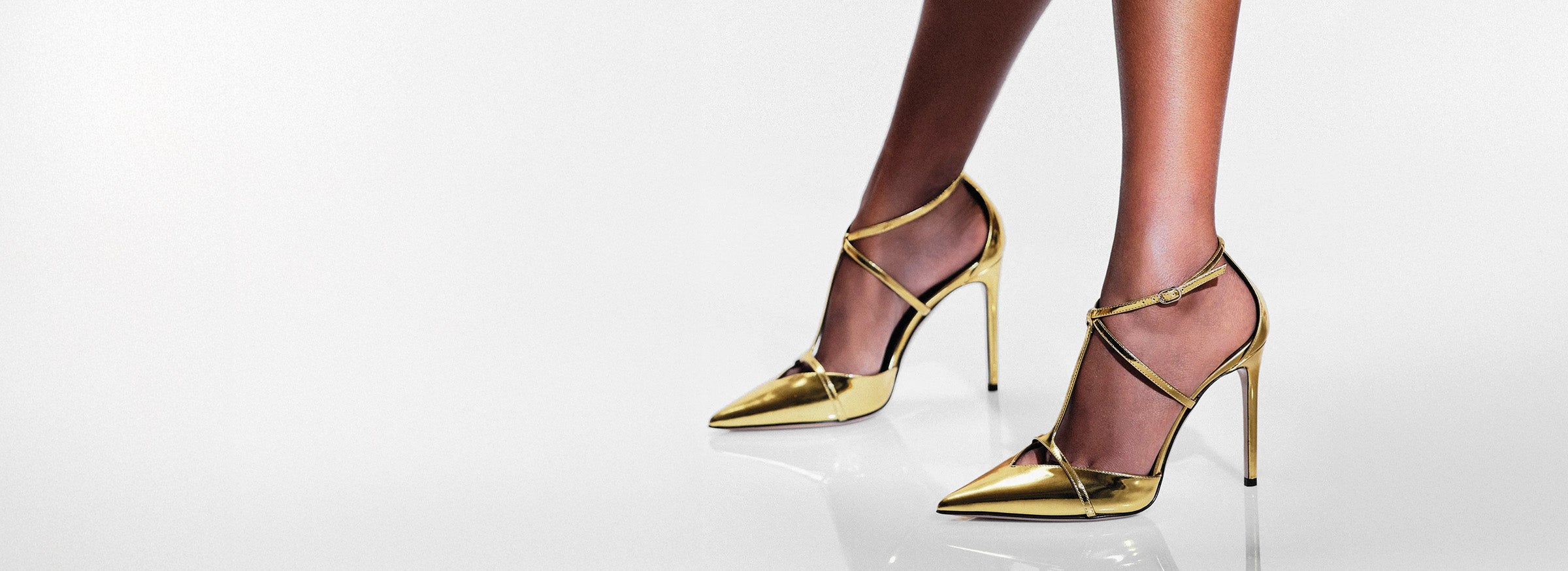  A pair of slender legs wearing shiny gold high heels with ankle straps on a reflective white surface, conveying elegance and glamour. Tap to shop