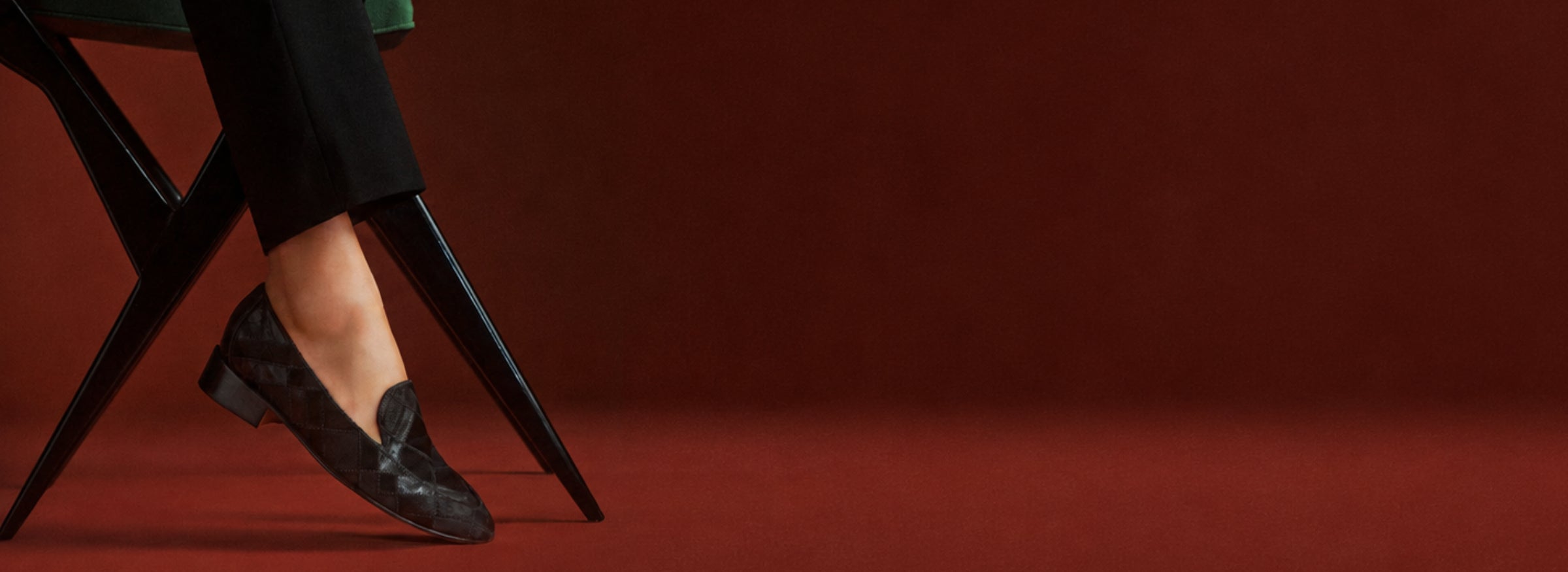  A seated person in cropped black trousers wears a dark woven leather loafer, legs crossed on a green folding chair against a warm rust-red backdrop; elegant, moody fashion editorial. Shop Now.