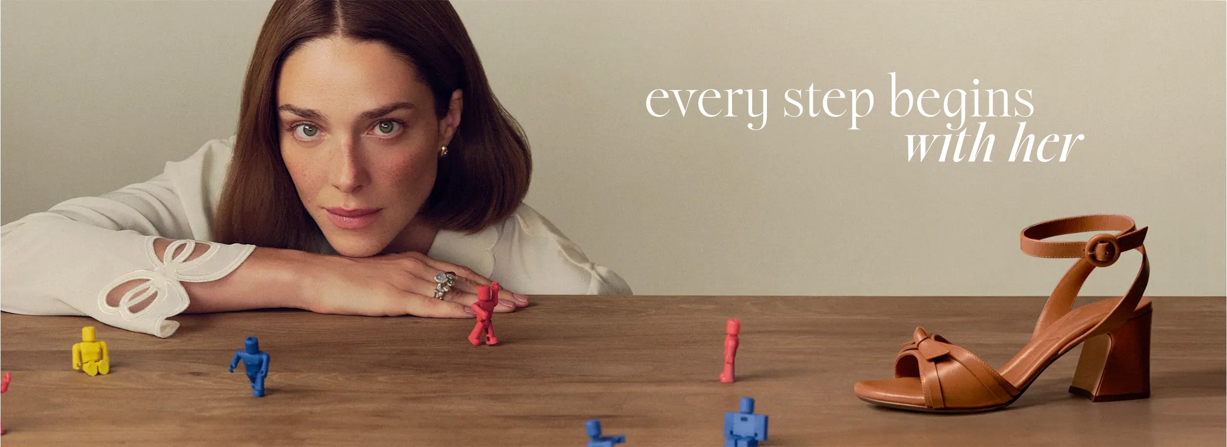  A woman in a white blouse gazes softly, her chin resting on a table with colorful toy figures. A brown sandal is on the right. Text reads "every step begins with her. Tap to shop