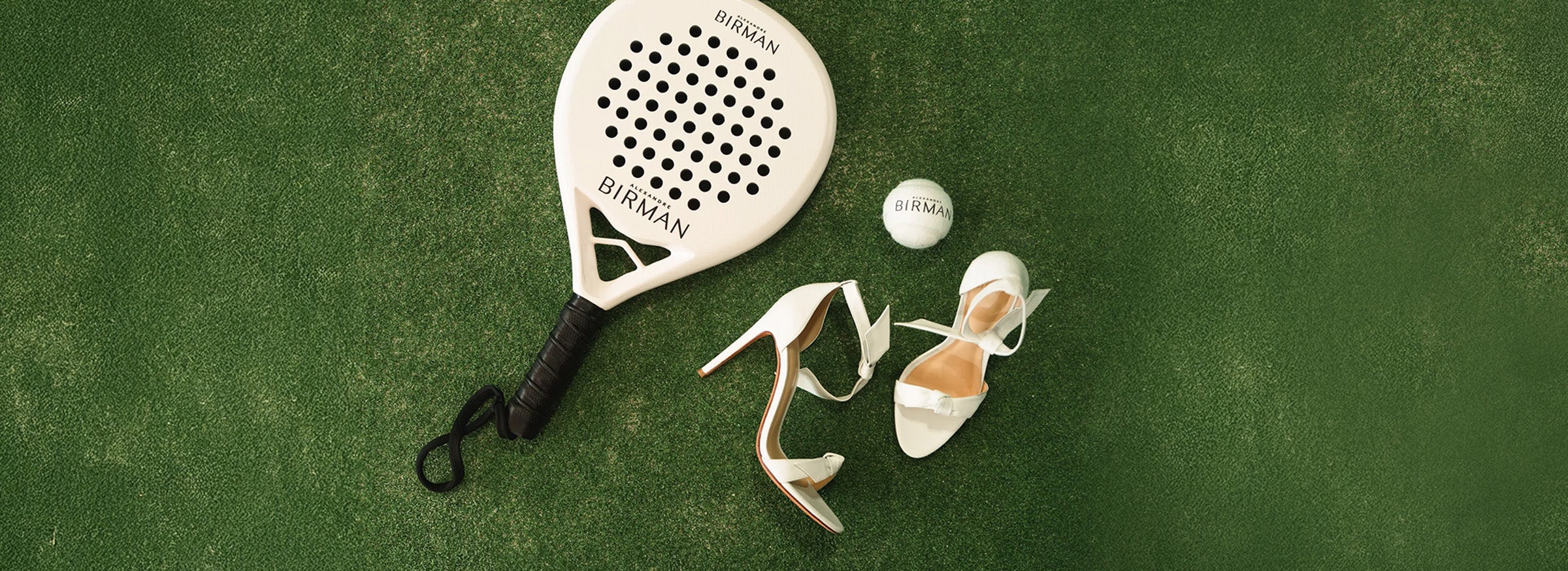  White Alexandre Birman Clarita sandals beside a branded padel racket and ball on a green court.