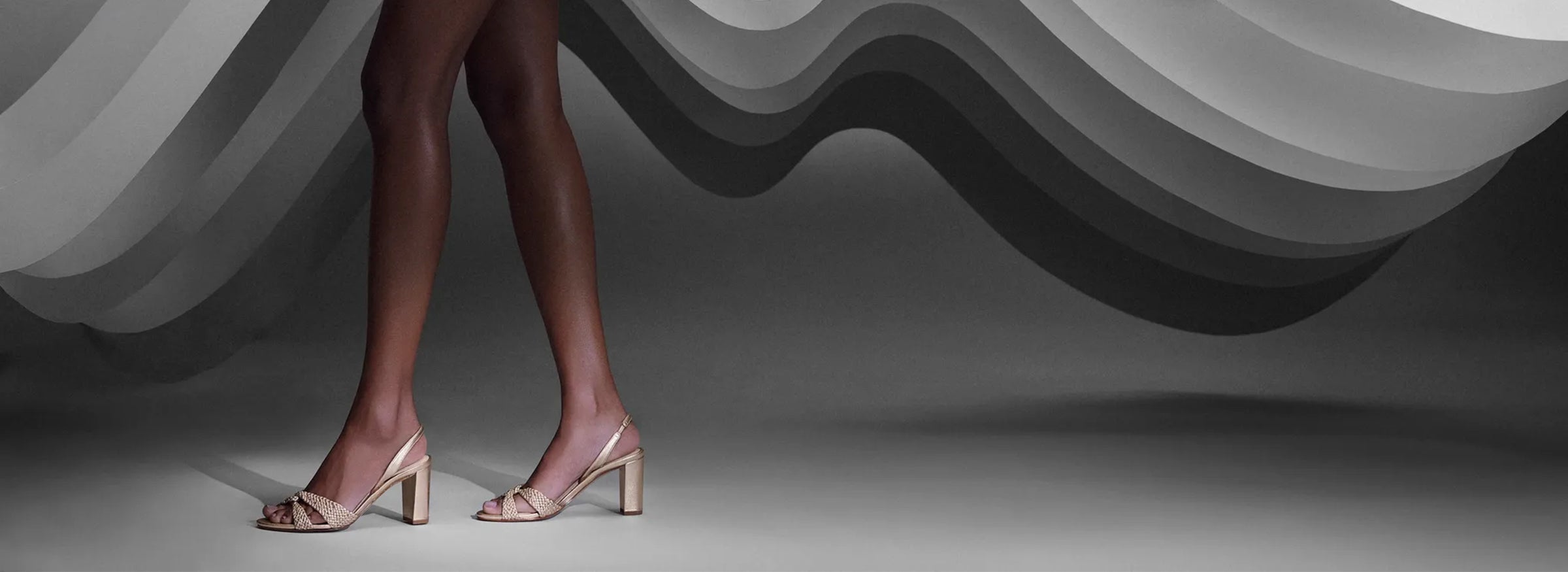  A model's legs in nude, open-toe heels stand gracefully on a gray background with wavy, layered patterns. The scene conveys elegance and sophistication. Tap to shop
