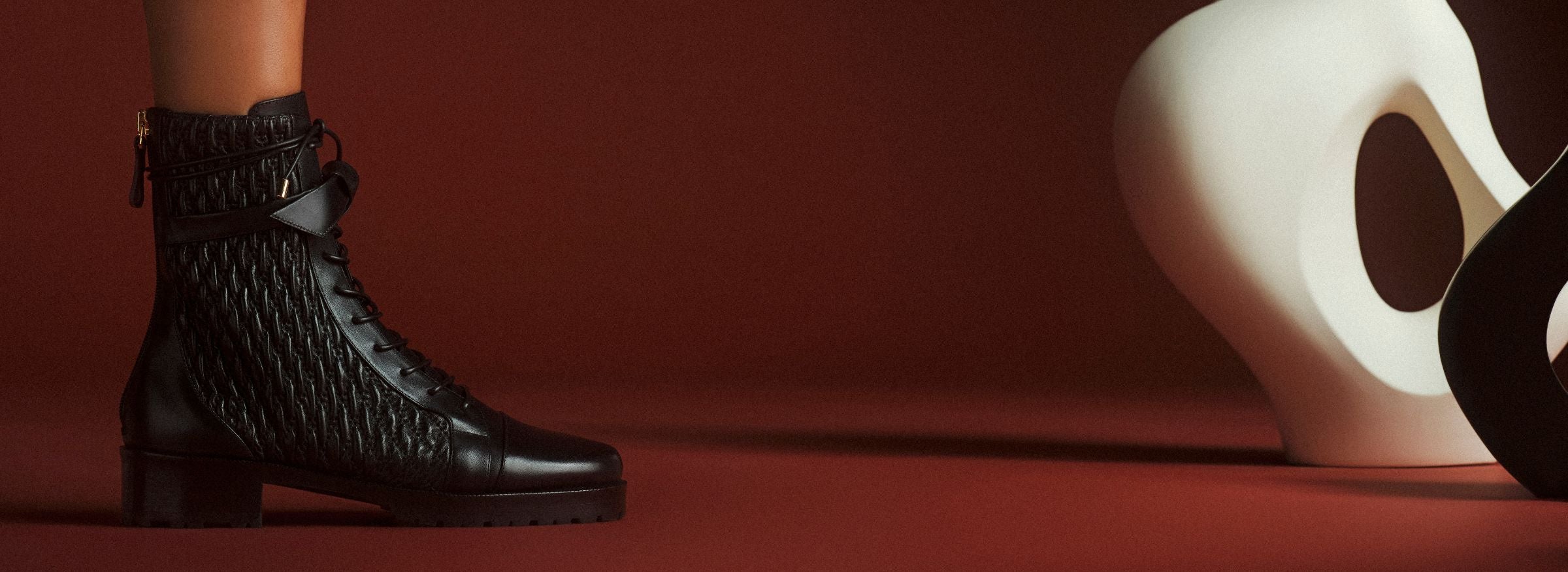  Close-up of a black textured ankle boot beside a white abstract sculpture on a deep red floor and backdrop, creating a bold, artistic mood. Shop Now.