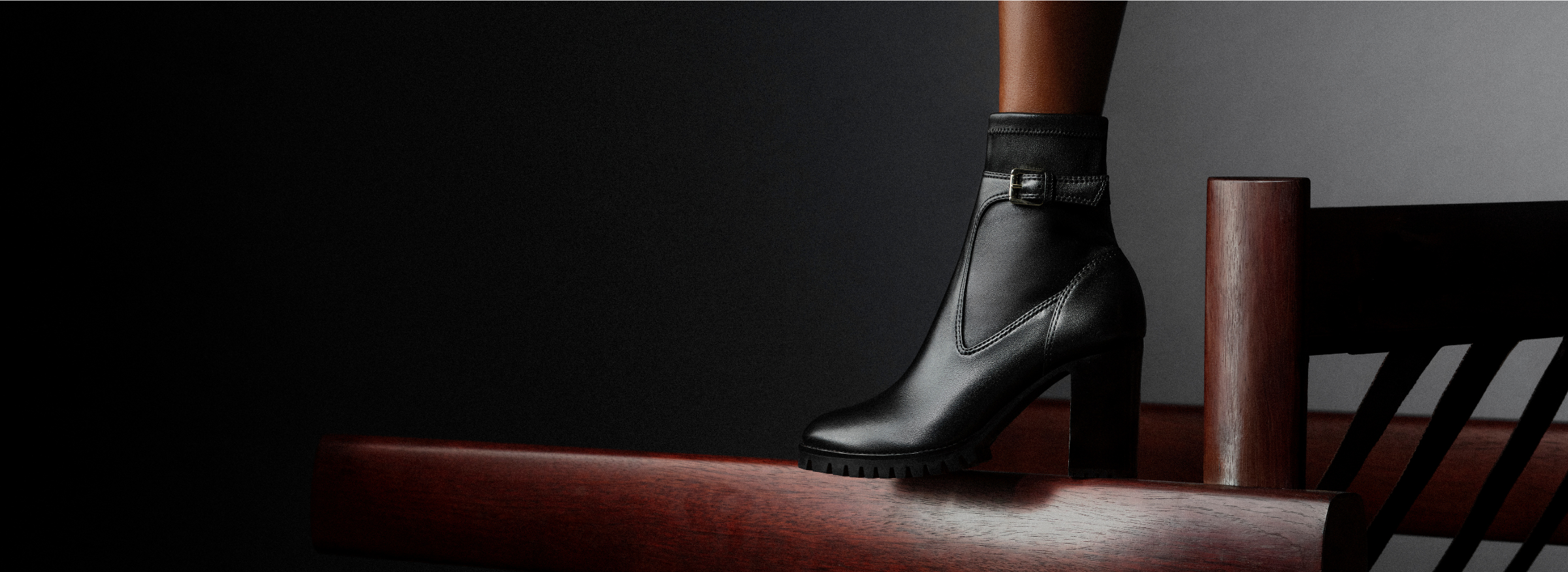  Black leather ankle boot with a high block heel rests on a wooden rail against a dark background. Shop Now.