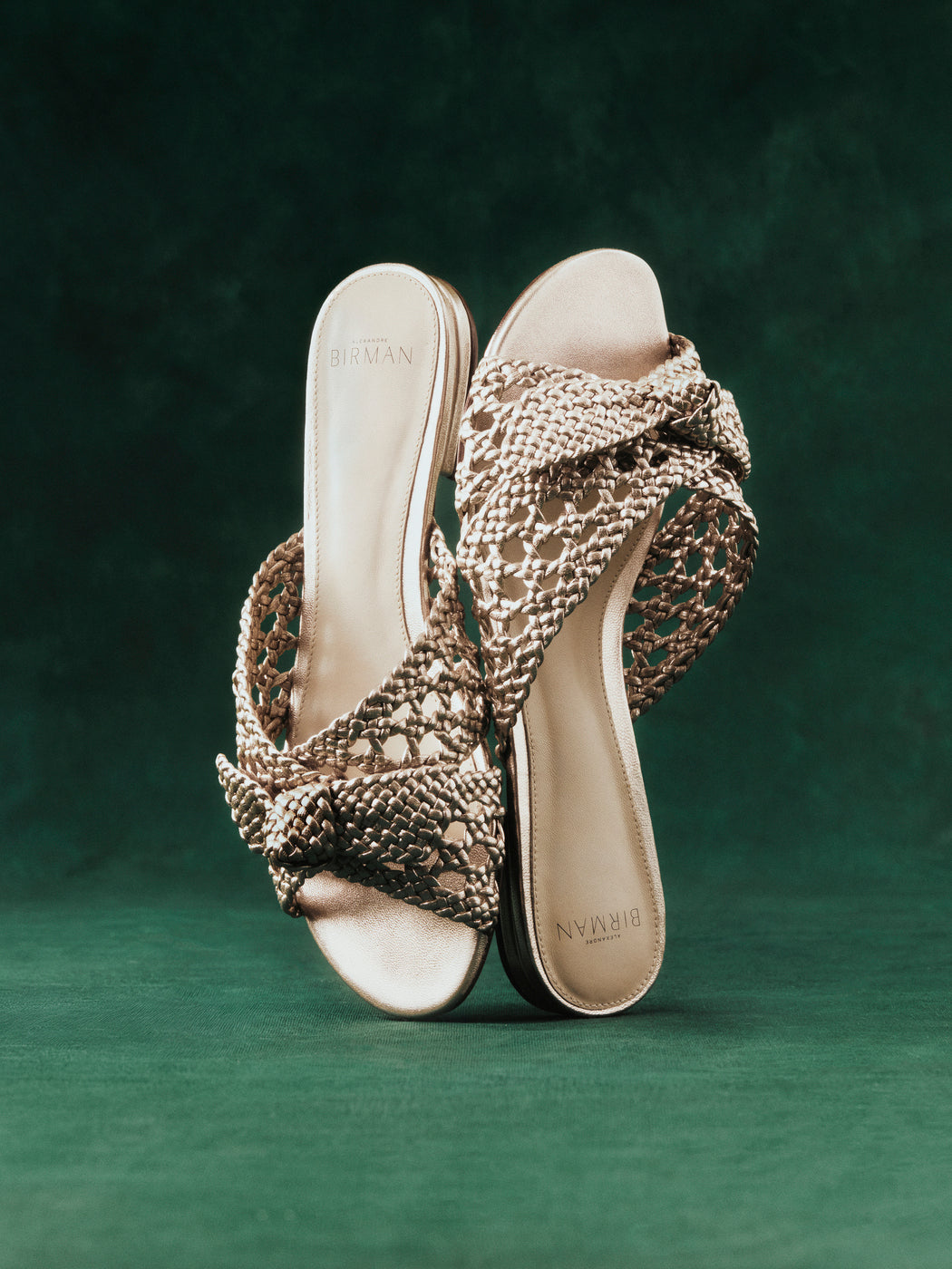 A pair of beige woven sandals with a bow design is displayed against a rich green background. The sandals are elegantly arranged upright. Tap to shop Flats