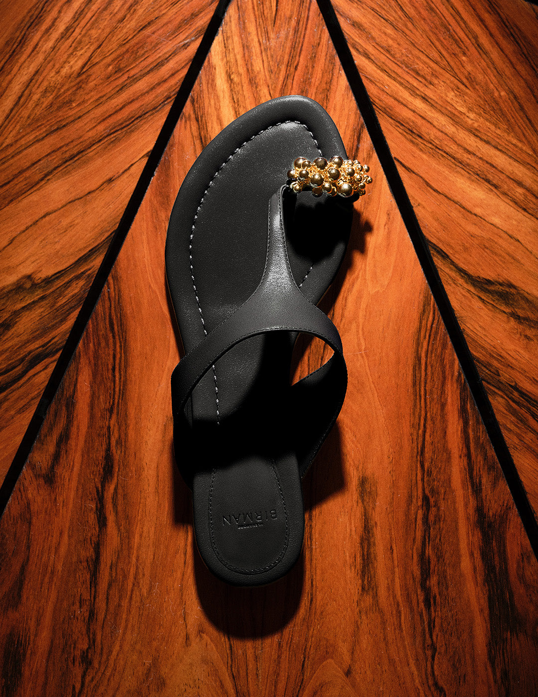 A top-down view of a black flat thong sandal resting on a geometric, wood-grain surface. The sandal is embellished with a cluster of gold spherical beads on the toe strap.