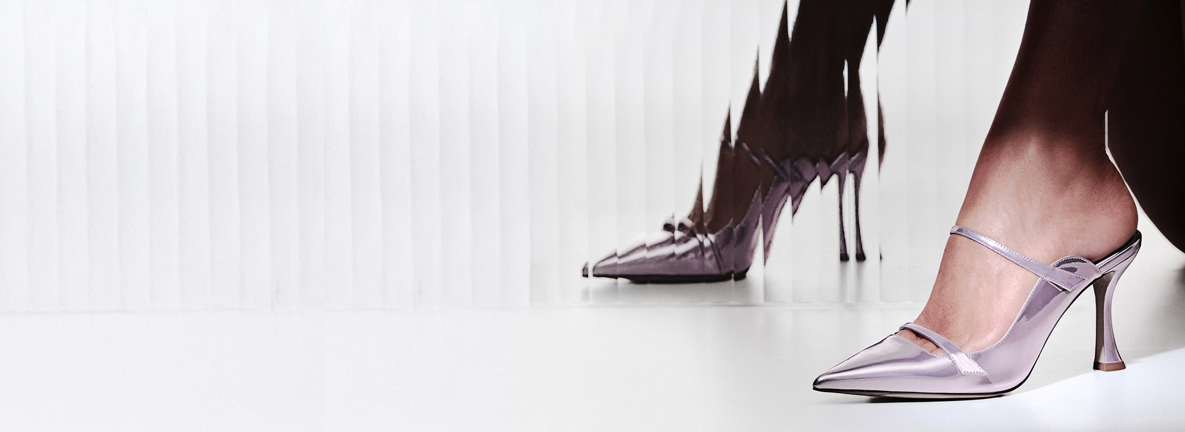  Metallic lavender pointed-toe mule with a thin, sculpted heel. The shoe features two slender straps across the instep. A model is seated next to a ribbed glass panel that creates a distorted reflection of the heels.