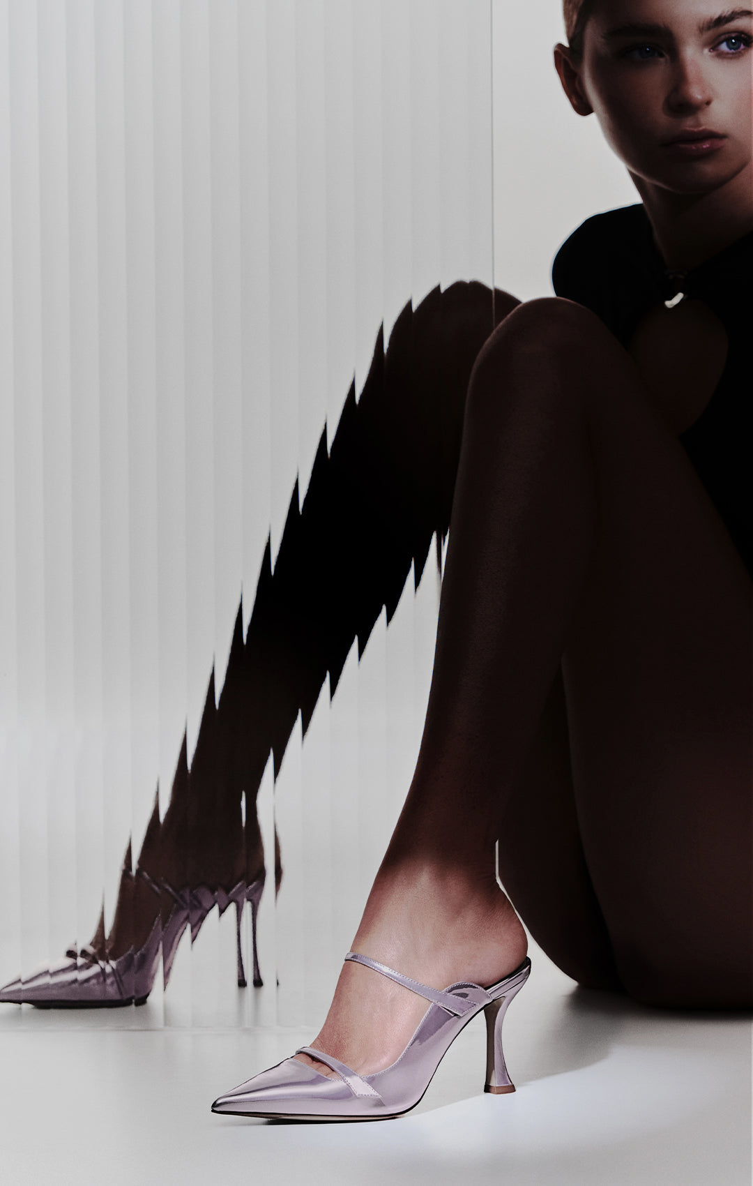  Metallic lavender pointed-toe mule with a thin, sculpted heel. The shoe features two slender straps across the instep. A model is seated next to a ribbed glass panel that creates a distorted reflection of the heels.