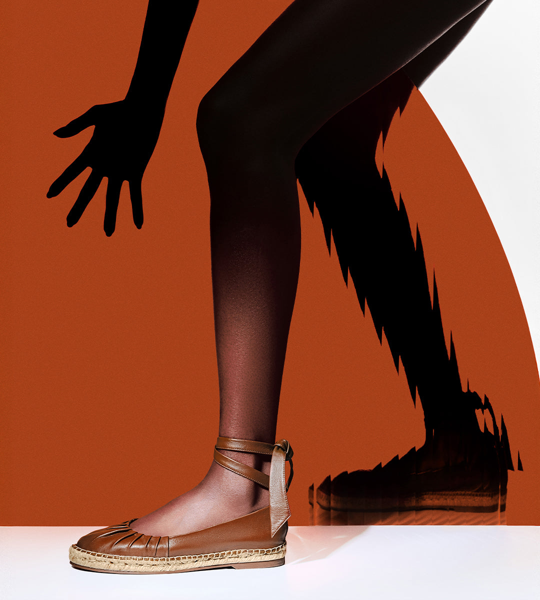 Brown leather flat shoe with an espadrille-style jute rope sole. The shoe has a gathered, ruched design at the toe and wraps around the ankle. It is worn by a model against a graphic orange and white background with artistic shadows.