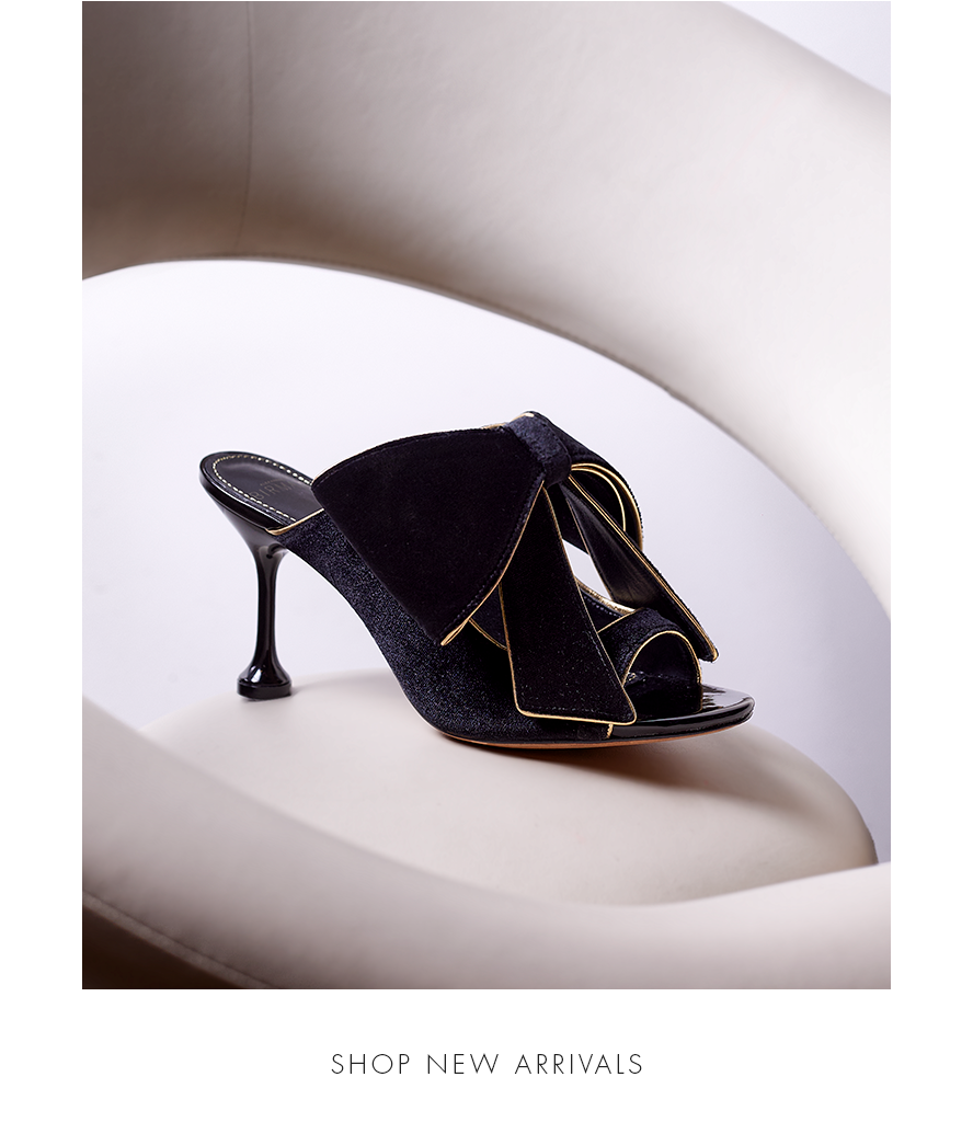 Close-up of the Luxurious Lupita Sandal in Black Velvet with a Stunning Bow Accent, Resting on a Modern White Chair. Text reads: SHOP NEW ARRIVALS