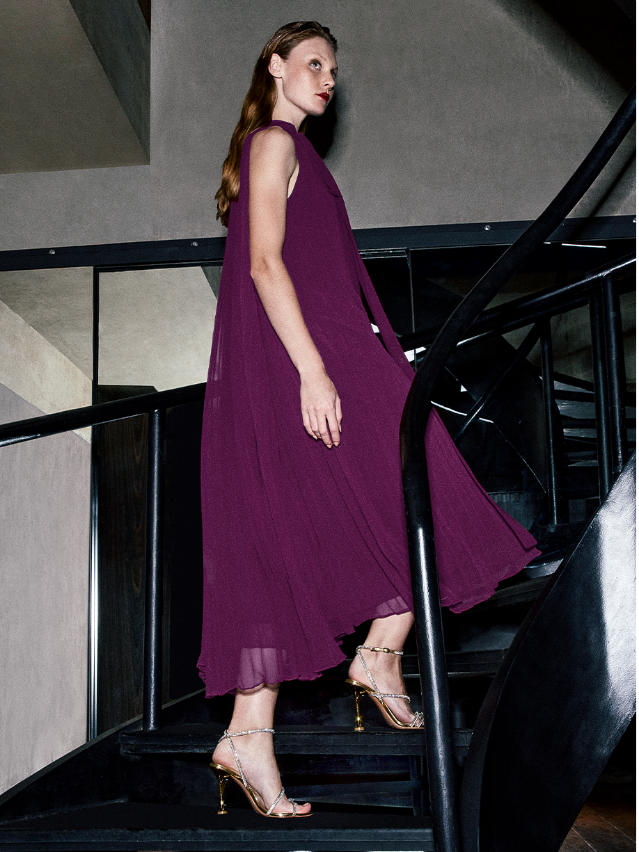 A woman climbing a chic staircase in a purple flowing gown. She showcases the Nina Sandal 100 in Oro, a stunning handcrafted sandal with zirconia embellishments.