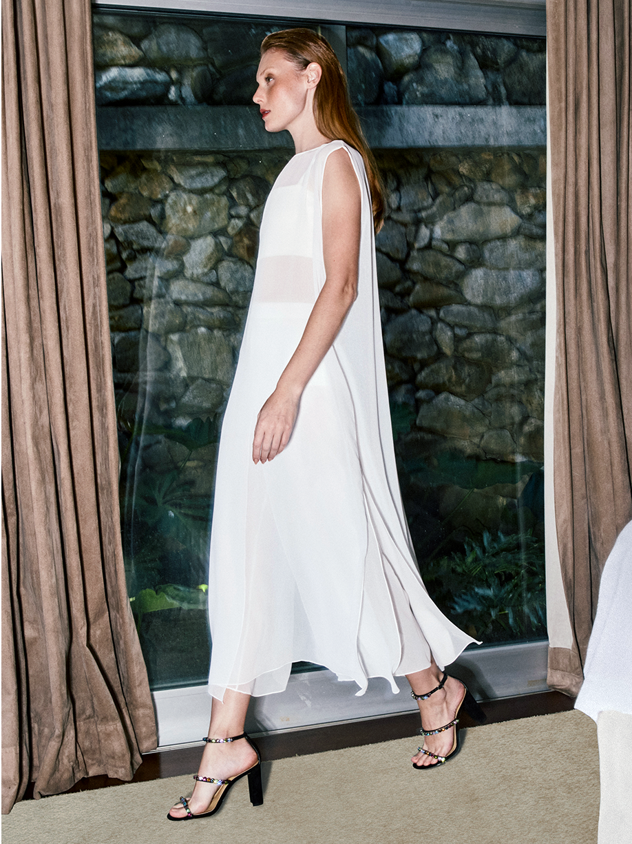 Crystal Heeled Beauty: A confident model stands in a room wearing a white flowy dress. She showcases the Alexa Crystals 90 Sandal with a striking black block heel.