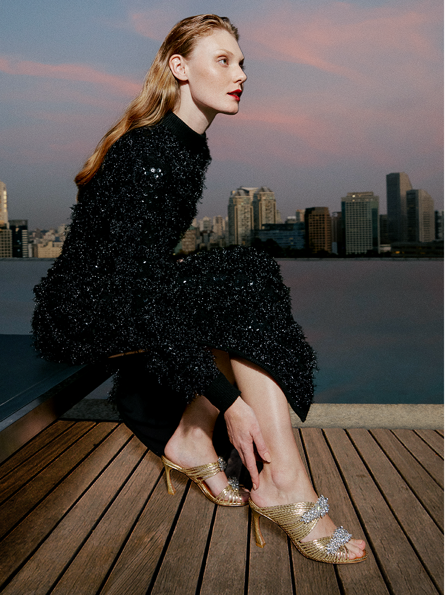 Golden Glamour: A woman sits on a bench with a stunning landscape behind her, wearing a black long dress embellished with sparkling sequins. Her hand touches the Lisa Crystal 85 Mule Oro, a luxurious gold-accented sandal.