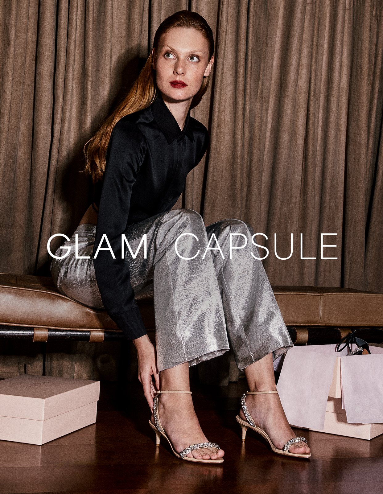 Luxury Glamour: A woman seated elegantly in a luxurious bedroom, wearing a black blouse and silver pants. She showcases the Aurora Crystal 50 Sandal in Beige, an embellished masterpiece.